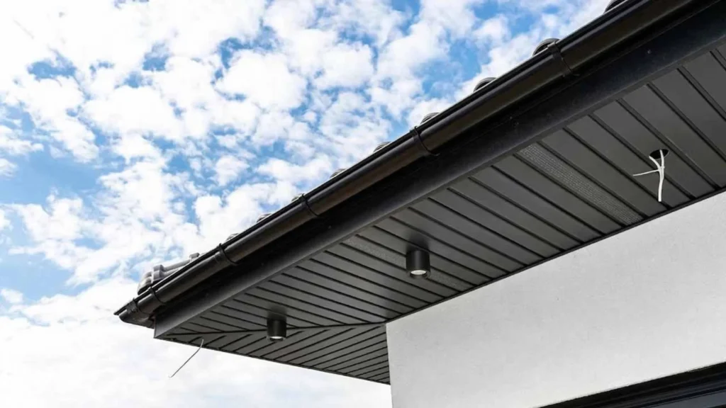 Close-up of a modern house's black soffit and gutter system against a bright blue sky with clouds. Clean lines convey a sleek, contemporary design.