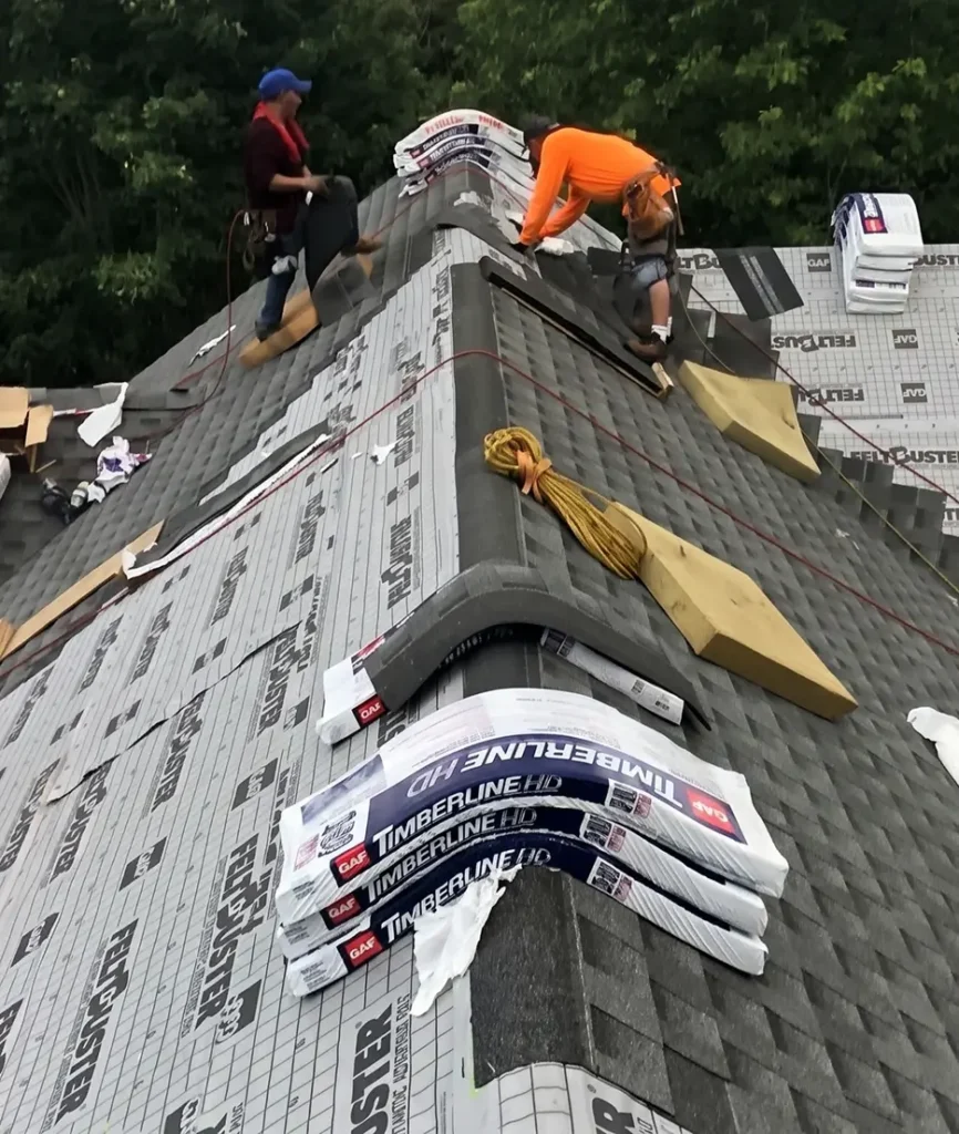 Roofers in work gear install shingles on a steep gray roof. Roofing materials and tools are scattered around. The scene is active and industrious.