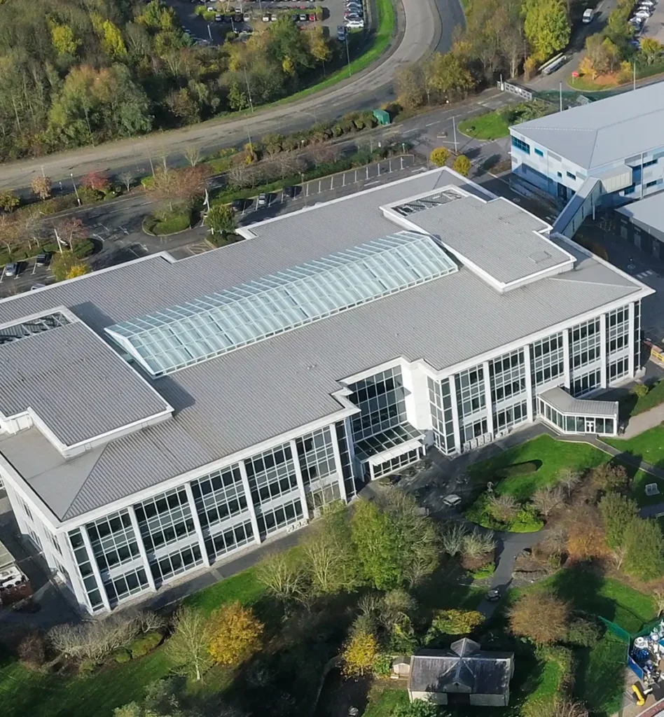 Aerial view of a large modern building with a glass-roofed atrium, surrounded by landscaped green areas and curved roads. The scene is calm and orderly.