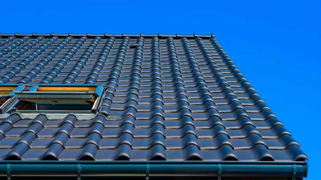 A tiled roof with a single skylight window under a clear blue sky. The shiny, dark tiles create a pattern, conveying a sense of structure and calm.