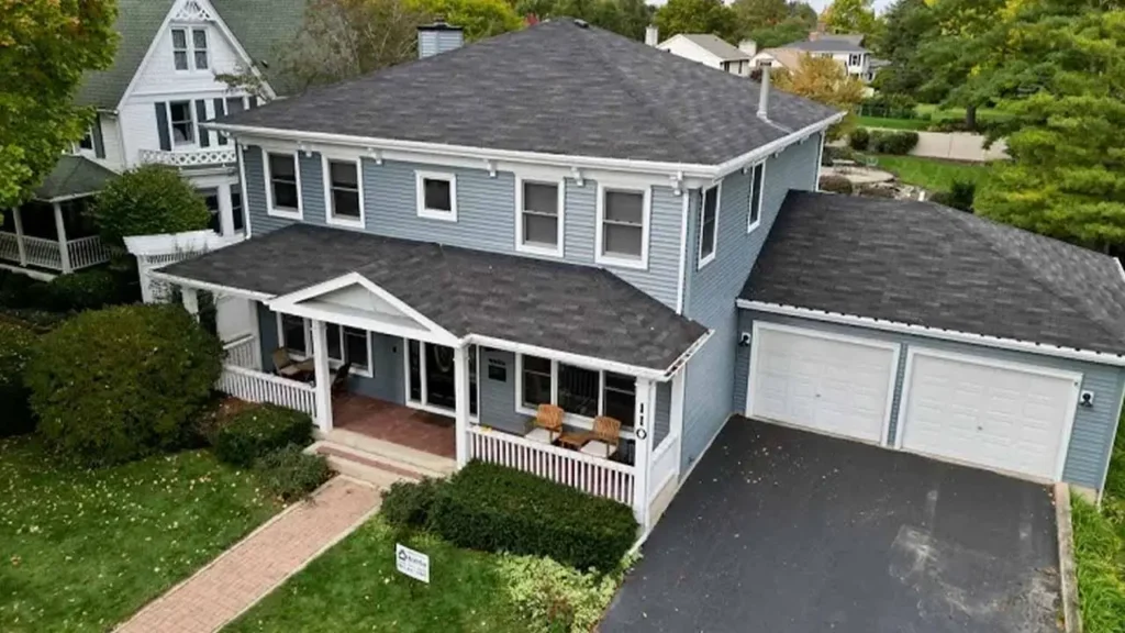 Aerial view of a two-story blue house with a front porch, white trim, and twin garages. Surrounded by greenery, it has a welcoming, suburban feel.