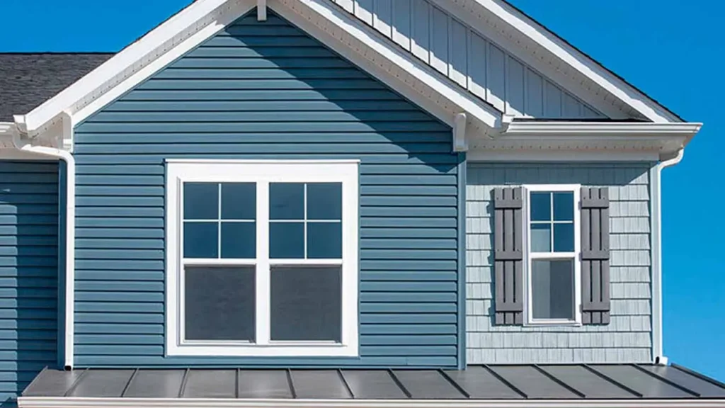 Close-up of a house exterior with blue siding, featuring a white-framed double window and gray shutters. Clear blue sky enhances the serene appearance.