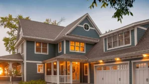 A cozy, modern two-story house with blue siding and warm, glowing windows at dusk. It features a gabled roof, white trim, and a covered porch.