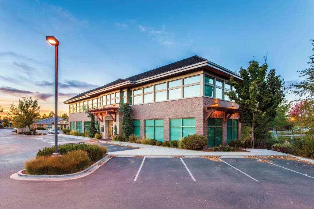 Modern brick-and-glass office building with teal windows beside an empty parking lot at sunset, surrounded by trees and landscaped shrubs.